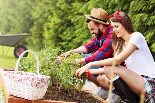 A man and a woman gardening outdoors in a backyard with lush green trees in the background. They are both crouching near a small raised flower bed, tending to leafy green plants with their hands. The woman is wearing a white t-shirt, a red headband, and plaid shorts, while the man is dressed in a red and blue checked shirt, a straw hat, and jeans. To the left, a white wicker basket with a floral pattern sits on the ground, containing gardening tools and supplies. The garden features neatly maintained soil, vibrant green plants, and a grassy lawn area in the foreground, indicating a well-kept residential space. The weather is sunny, with bright natural light illuminating the scene, reflecting typical outdoor gardening activities associated with local landscaping services in Hoxton or nearby London areas.