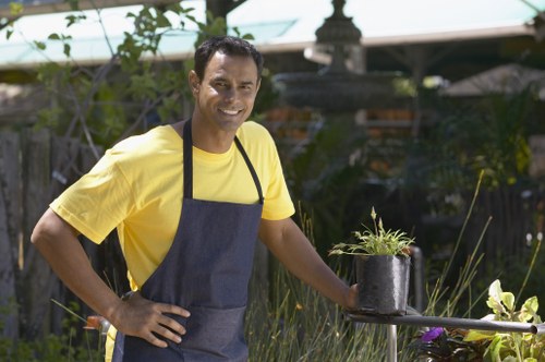 A man with medium skin tone wearing a yellow T-shirt and a dark grey apron stands in a well-maintained garden, smiling at the camera. In his right hand, he holds a small black pot containing a green, leafy plant, and his left hand rests on his hip. The garden features a neatly edged lawn with dense, green grass in the foreground, bordered by an array of shrubbery, flowering plants, and small bushes. Behind him, there are taller plants and a wooden fence providing privacy, with some garden tools visible on a surface nearby. The background also shows garden structures, including a greenhouse with a pale green roof and a decorative stone fountain or birdbath. The scene is illuminated by natural daylight, suggesting a clear weather day, with soft shadows and vibrant plant colours typical of a professional gardening or landscaping service environment in Hoxton or nearby London. This image aligns with gardening and outdoor maintenance services offered by Landscapers Hoxton.
