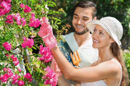 A woman and a man working together in a lush, well-maintained garden with vibrant pink flowering bushes, green foliage, and a neatly shaped hedge in the background. The woman, wearing a white sleeveless top, a wide-brimmed hat, and gardening gloves, is carefully pruning or tending to the plants with a garden tool, while the man, dressed casually and smiling, assists and observes. The garden has a mix of flowering plants, green grass, and likely other garden features such as soil and paving visible in the scene. Bright natural daylight illuminates the outdoor space, suggesting a clear, sunny day, ideal for outdoor gardening activities. The scene reflects typical lawn and garden care practices, and indicates professional gardening or landscaping maintenance that a company like Landscapers Hoxton might provide in the local area of Hoxton or Greater London.