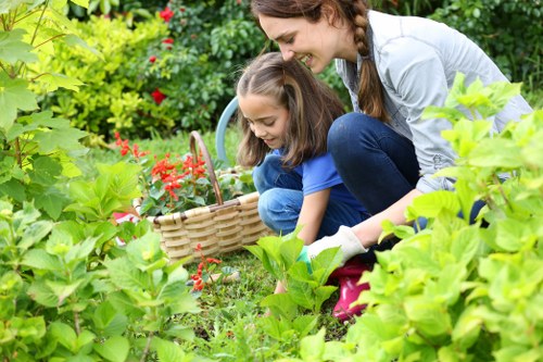 A young woman with long brown hair and a girl with shoulder-length hair are gardening together in a lush outdoor space, surrounded by vibrant green foliage, flowering plants, and well-maintained grass. The woman is crouched down, smiling kindly as she assists the girl in selecting or planting flowers from a woven basket filled with small red blossoms. The garden features a variety of plants, including hedges, flowering shrubs, and leafy bushes, with a backdrop of dense greenery. The ground is covered with neatly trimmed grass, and the overall environment appears to be a well-kept, landscaped garden suitable for outdoor family activities and gardening projects. The scene is lit by natural daylight, suggesting a clear day, perfect for gardening tasks. This natural, welcoming setting underscores the importance of outdoor maintenance and gardening services offered by companies like landscapershoxton.co.uk, highlighting their expertise in creating and maintaining beautiful garden environments in Hoxton and surrounding areas.