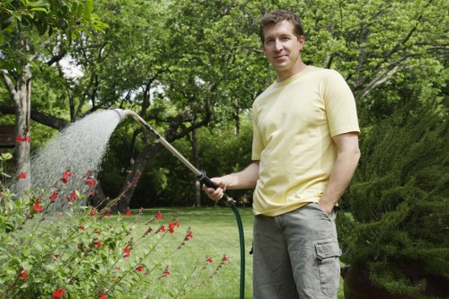 A man with short brown hair and a light yellow t-shirt stands in a well-maintained private garden, holding a garden hose that is spraying water onto a lush flower bed with small red flowers and green foliage. The garden features a neatly mowed lawn with vibrant green grass, bordered by a hedge on one side and mature trees with dense green leaves providing shade in the background. The scene appears to be during daytime with natural sunlight illuminating the garden, creating a peaceful outdoor environment suitable for gardening and lawn care services offered by Landscapers Hoxton, situated near the postcode area of N1 in London, supporting sustainable gardening practices with a focus on environmentally conscious outdoor maintenance.