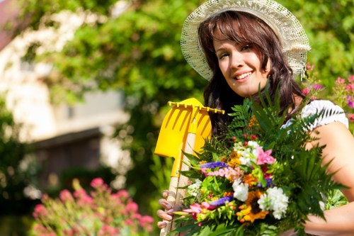 A woman in a garden during daylight hours, wearing a wide-brimmed straw hat and holding a colorful bouquet of mixed flowers, greenery, and garden foliage. In the background, there is a lawn with lush green grass, bordered by a hedge and various plants, with some flowering shrubs visible. The garden features a neatly maintained lawn, with a paved pathway or patio area partially visible in the foreground, and trees with bright green leaves providing dappled sunlight. The scene captures a vibrant outdoor space typical of a landscaped garden in a residential area near Hoxton, with natural tones of green, pink, and white, and hints of purple and yellow from the flowers. This image reflects outdoor gardening and floral arrangement activities that landscaping services like those from Landscapers Hoxton often support, showing a well-kept and inviting garden environment under soft, natural lighting.