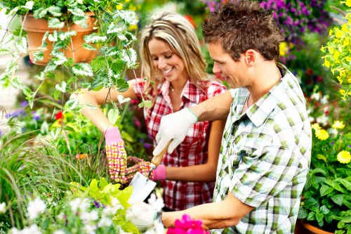 The image depicts a brightly lit garden scene with a woman and a man engaged in gardening activities amidst lush greenery and vibrant flowering plants. The woman, wearing a red plaid shirt and pink gardening gloves, is smiling as she tends to a plant, holding pruning shears in her right hand. The man, dressed in a light-colored, checked short-sleeve shirt, is trimming or examining a shrub with a garden tool in his right hand. The garden features a well-maintained lawn in the foreground, with dense foliage including flowering plants with purple, yellow, and pink blooms, and neatly edged flower beds. In the background, there are tall, leafy trees and a mix of broad-leafed shrubs, creating a rich, natural environment typical of a landscaped outdoor space in Hoxton, London. The scene suggests outdoor gardening and lawn care, supported by a local landscaping company, with natural daylight highlighting the healthy, thriving plants and the vibrant colours of the flowers and foliage, all set under a partly cloudy sky typical of a mild weather day. This detailed scene exemplifies professional garden maintenance and landscaping services in an urban outdoor setting.