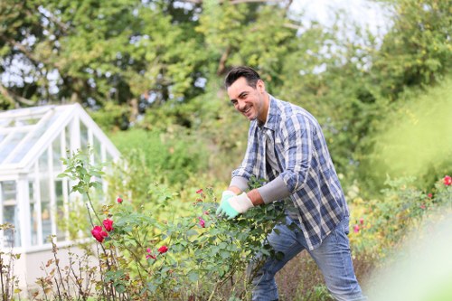 A man engaged in gardening within a well-maintained backyard garden, surrounded by various flowering plants and shrubs. The garden features a lush green lawn area and bordered flower beds with pink, red, and purple blooms, indicating a diverse planting scheme. In the background, there are mature trees and a small greenhouse structure, suggesting a productive outdoor space. The man is wearing casual clothing, including a blue plaid shirt and jeans, and is smiling while tending to the plants, possibly pruning or planting. The overall scene is set during daylight with natural lighting, and the environment appears tidy and cultivated, reflecting professional gardening care that could be offered by landscapers such as Landscapers Hoxton. This setting highlights the relevance of routine garden maintenance, plant care, and outdoor landscaping services in the Hoxton area, contributing to a vibrant and inviting outdoor space.