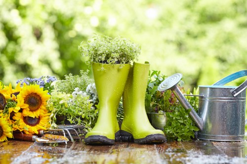 A garden scene featuring a weathered wooden table with various gardening tools and decorative elements. In the foreground, a pair of bright green rubber rain boots filled with small white flowers serve as planters. To the right, a silver metal watering can with a blue handle sits next to a yellow-handled trowel, both on the table. On the left, a bunch of vibrant yellow sunflowers and other colourful blooms rest beside a pair of pruning shears. The background shows a lush, green garden with blurred foliage, suggesting a well-maintained outdoor space that could be in Hoxton or a similar London neighbourhood. The lighting indicates a bright, possibly sunny day, emphasizing the natural tones and textures of the garden elements and tools. This image captures the essence of outdoor gardening activities and encourages professional garden maintenance and landscaping services offered by Landscapers Hoxton, focusing on garden beautification and lawn care in the local area.