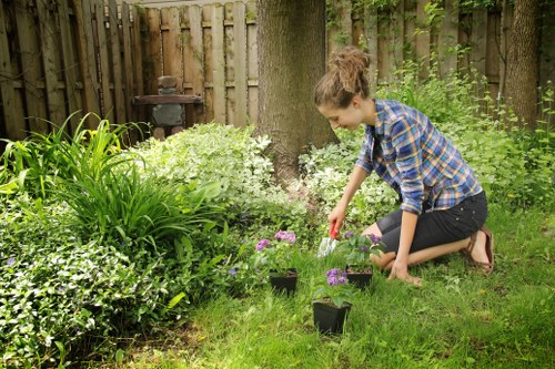 A young woman with curly brown hair, dressed in a blue plaid shirt and dark jeans, kneeling on a well-maintained grass lawn in a back garden in Hackney, London, surrounded by lush green plants, shrubs, and small flowering plants in purple and white. She is watering newly planted flowers in black plastic pots using a small watering can, with a wooden picket fence and mature trees visible in the background. The garden features a mix of natural grass, diverse foliage, and paved area, creating an inviting outdoor space suitable for gardening and landscaping services by Landscapers Hoxton.
