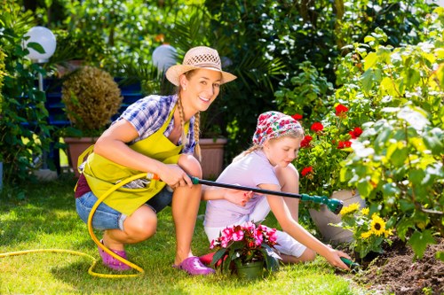 A woman and a young girl are gardening together in a lush backyard, surrounded by vibrant green foliage, shrubs, and flowering plants. The woman, wearing a straw hat, yellow apron, and checkered shirt, is crouched down and holding a garden hose, smiling at the camera. The girl, dressed in a light-colored dress and a colorful bandana, is also crouched, closely tending to a flower bed with pink and yellow blooms, using her hands to soil the plants. The garden features a well-maintained grassy lawn in the foreground, with a paved pathway visible near the flower beds. In the background, there are dense hedges, trees, and hanging garden lights, indicating a private, landscaped outdoor space typical of a personal garden in Hoxton. The scene suggests an outdoor activity in pleasant weather, ideal for lawn and garden maintenance as provided by local gardening services. Overall, the image captures a cheerful moment of outdoor gardening in a vibrant, well-kept garden environment.