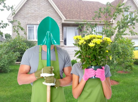In a well-maintained front garden with lush green grass and a paved pathway, two individuals are standing side by side, both wearing green gardening aprons and gloves. The person on the left is holding a large garden spade with a turquoise handle, while the person on the right is holding a vibrant pink flower pot filled with yellow flowering plants. Behind them, a brick house with white-framed windows and a grey tiled roof is partially visible, framed by bushes and small trees, some with light green leaves. The garden features a neatly edged lawn, a variety of shrubs, and a few young trees, suggesting a healthy and cared-for outdoor space. Soft, natural daylight indicates a clear day, ideal for gardening. The scene emphasizes outdoor garden maintenance and planting activities typical of professional gardening services offered by Landscapers Hoxton in the Hoxton area, touching on landscape enhancement, flowerbed planting, and lawn care within a suburban environment.