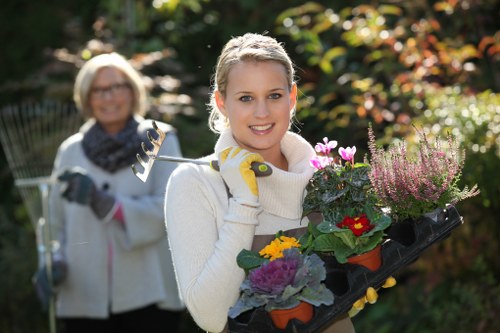 A young woman in a cream sweater and gardening gloves is smiling and holding a black tray with potted flowering plants, including red, purple, yellow, and pink blooms, in a well-maintained backyard garden during daylight. The garden features a lush, green lawn with neatly edged borders, a dense hedge in the background, and a variety of trees and shrubs providing a natural backdrop. The sunlight filters through the foliage, creating a warm and inviting outdoor environment, appropriate for gardening and landscaping activities. Behind the woman, an older woman wearing glasses, a white coat, and a blue scarf is partly visible, holding gardening tools and engaging in outdoor garden maintenance, reflecting typical gardening services offered by Landscapers Hoxton in the Hoxton area across London postcode regions. The scene captures a moment of gardening preparation or planting, emphasizing outdoor lawn care, flower bed planting, and landscape enhancement, all within a residential garden setting, suitable for homeowners seeking professional gardening services in the Hoxton vicinity.