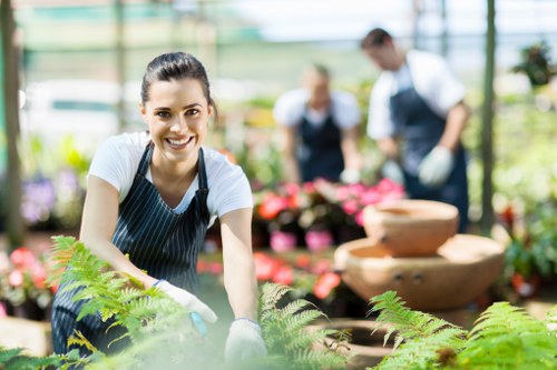 A smiling woman wearing a striped apron and gardening gloves is working in a greenhouse or garden centre with a variety of plants and flowers around her. In the background, two other people are seen tending to potted plants, suggesting outdoor gardening or landscaping activities. The area includes lush green foliage, vibrant pink flowers, and wooden decorative features such as a tiered garden fountain, all illuminated by natural daylight. The scene is set in a well-maintained garden or outdoor plant nursery, with visible glass walls letting in ample sunlight, highlighting the different textures of soil, foliage, and flowering plants. The overall environment displays a professional yet welcoming atmosphere consistent with gardening and landscaping services, suitable for inclusion on a site like landscapershoxton.co.uk, particularly within a page about planting services in the Hoxton area or related London districts.
